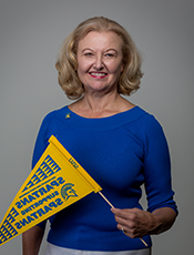 waist-up photo of Marcia Daszko, Caucasian female with medium length blonde hair, wearing blue long sleeved blouse with sleeves pushed up to ellbows, holding a yellow and blue 菠菜网lol正规平台 Spartan flag