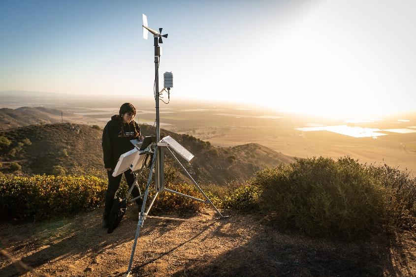 A students stands at the top of a high mountain while working on equipment to track wildfires.